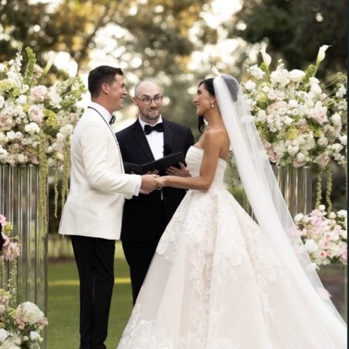 Bride and groom hold hands facing each other as an officiant reads vows outdoors among white and pink florals.