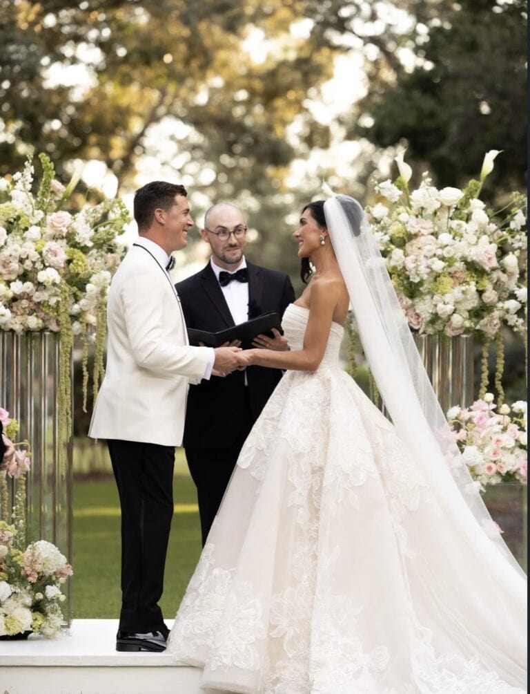 Screenshot | National Tuxedo Rentals Bride and groom hold hands facing each other as an officiant reads vows outdoors among white and pink florals.