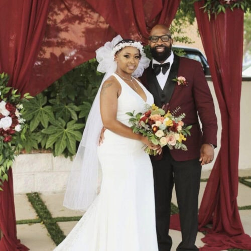 Bride in a white wedding gown and veil stands beside a groom in a burgundy suit under a maroon draped arch, holding a colorful bouquet for a wedding portrait.