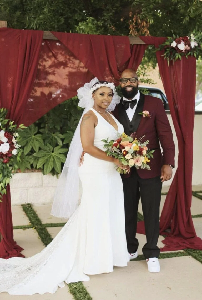 Bride in a white wedding gown and veil stands beside a groom in a burgundy suit under a maroon draped arch, holding a colorful bouquet for a wedding portrait.