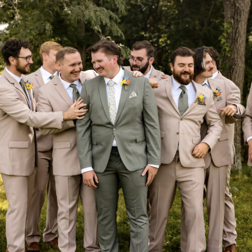 Group of men in beige suits outdoors, with a man in a green suit at the center smiling as friends surround him, boutonnières visible on lapels.