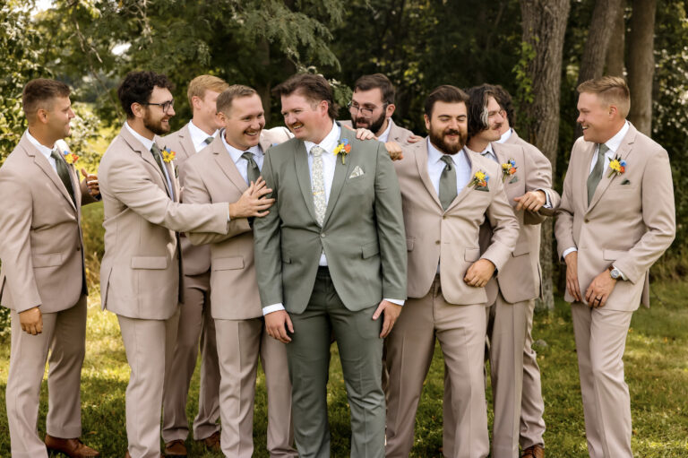 Group of men in beige suits outdoors, with a man in a green suit at the center smiling as friends surround him, boutonnières visible on lapels.