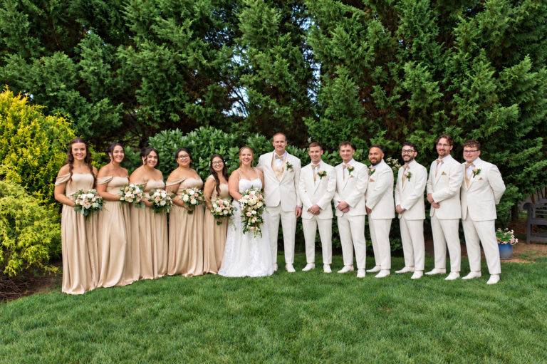 Bride and groom with bridesmaids and groomsmen posing on a green lawn in front of tall hedges; bridesmaids in champagne gowns, bride in white lace dress, groomsmen in cream suits with pale ties, all holding bouquets and boutonnieres.