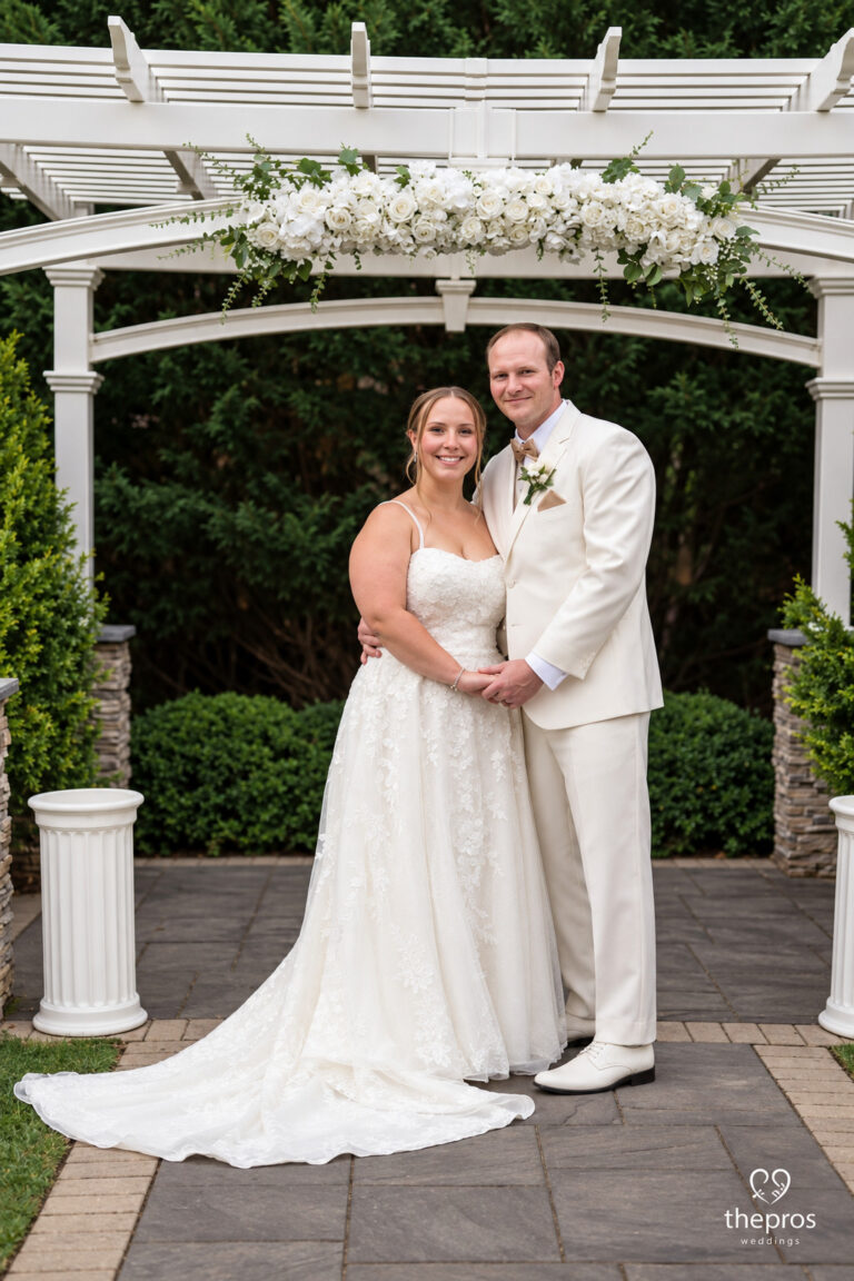 Bride and groom pose together under a white floral arch in a garden setting; she wears a lace wedding gown, he a white suit.