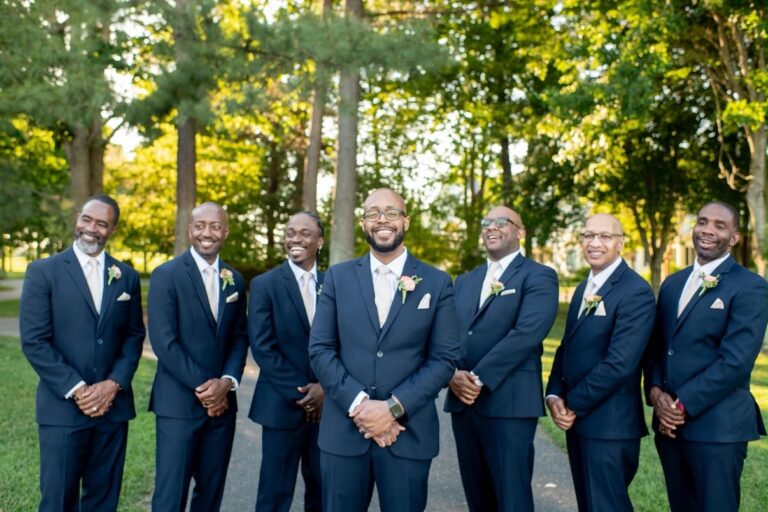 Seven men in navy suits with boutonnieres pose together on a sunny day in a park; central figure likely the groom.