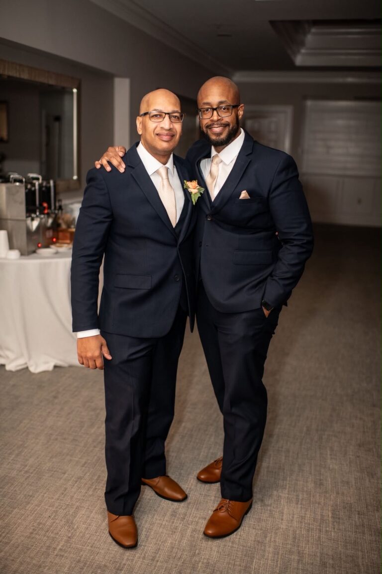 Two men in navy suits with beige ties smile and stand arm-in-arm at a formal event indoors.