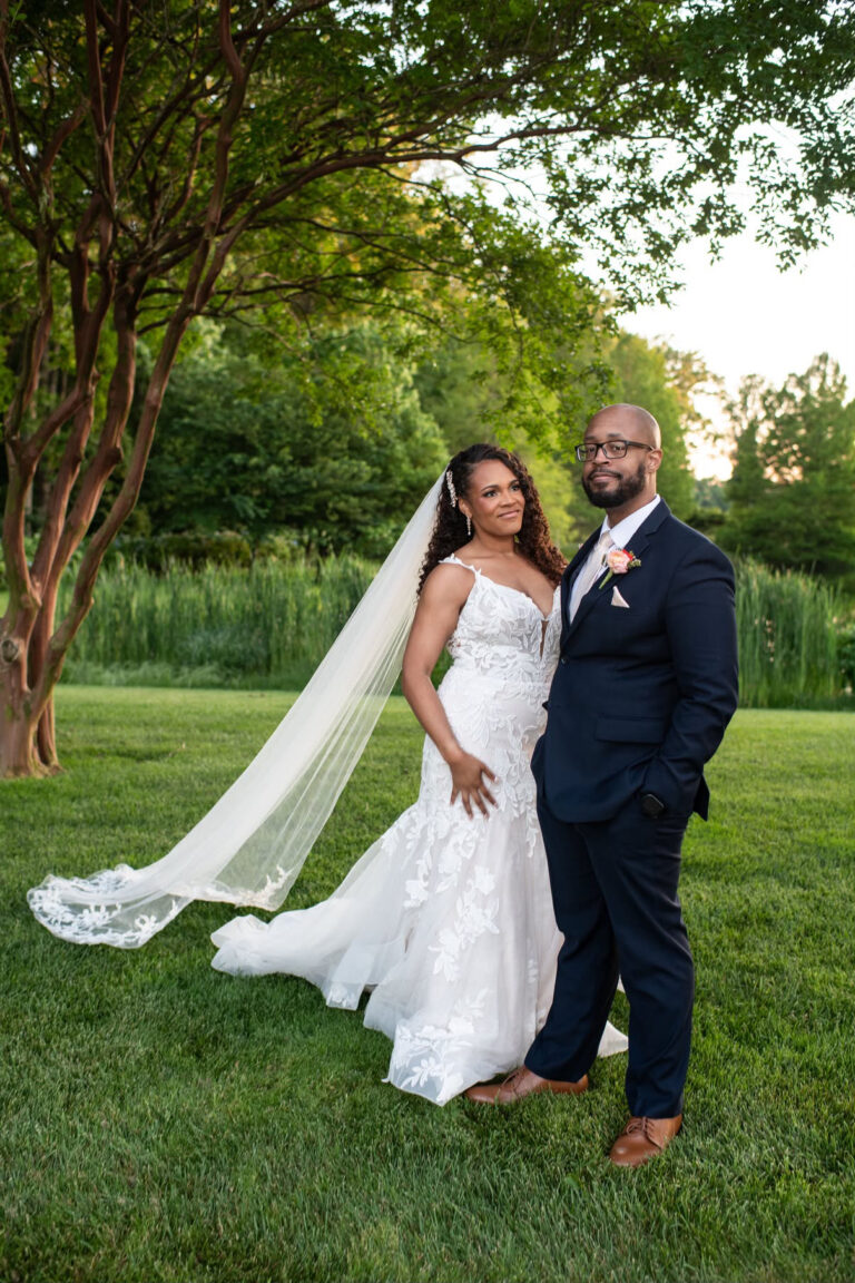 Bride in a white lace wedding gown with a long veil standing beside a groom in a navy suit on a green lawn under trees.