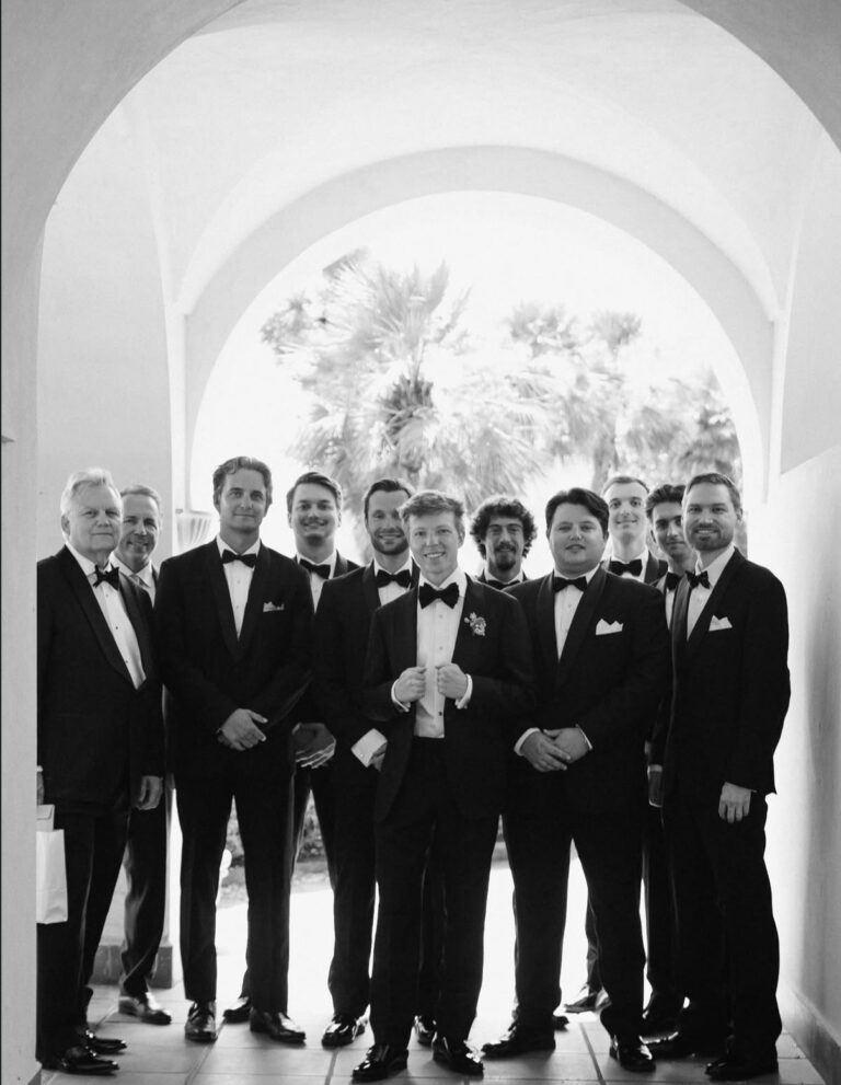 Groom and his groomsmen in black tuxedos posing under a white arch at a wedding.