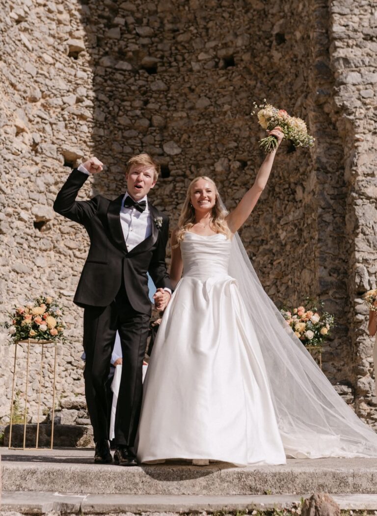 Bride in a white gown and groom in a black tuxedo celebrate on stone steps, bride raising a bouquet high above her head.