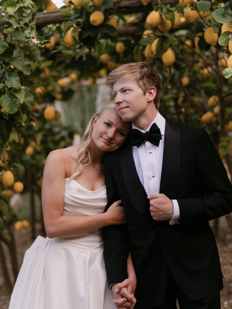 Bride in a white wedding dress leans her head on a groom in a black tuxedo, standing hand in hand beneath hanging lemon trees in a sunny garden setting.