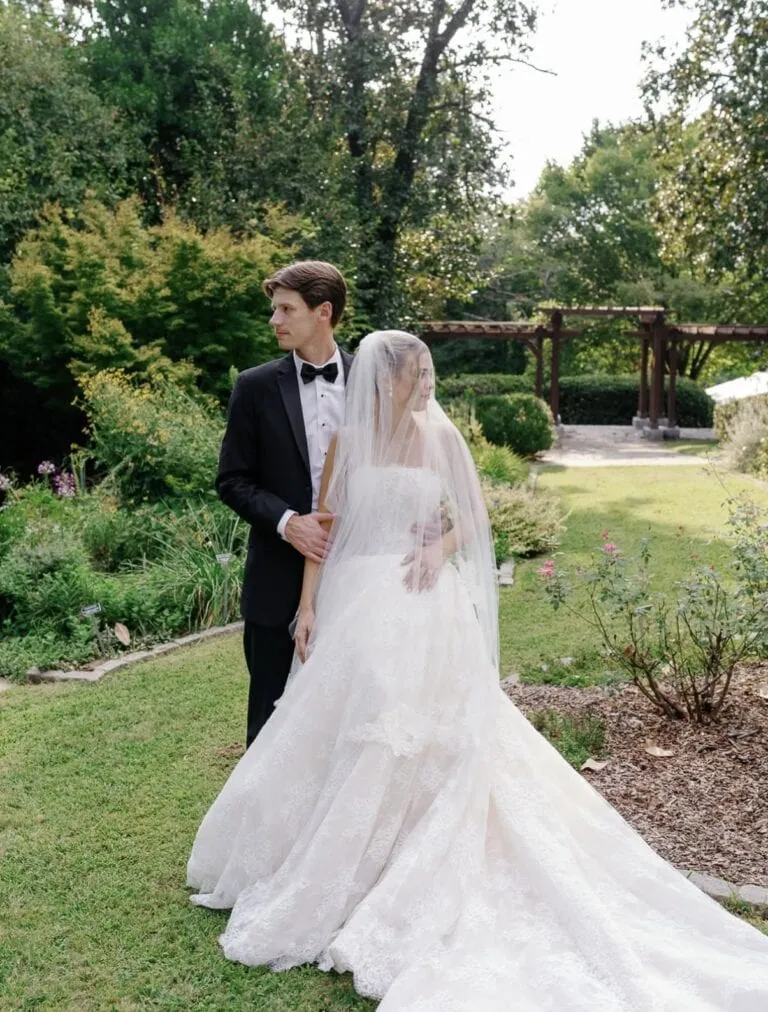 Screenshot | National Tuxedo Rentals Bride in a white wedding gown and veil stands beside a groom in a black tuxedo in a lush garden setting.