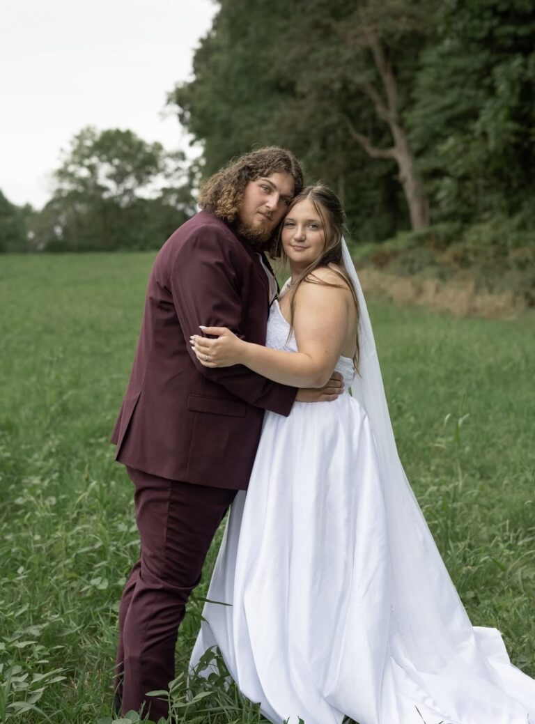 Bride and groom embracing in a grassy field with trees in the background, she in a white gown and veil, he in a burgundy suit.