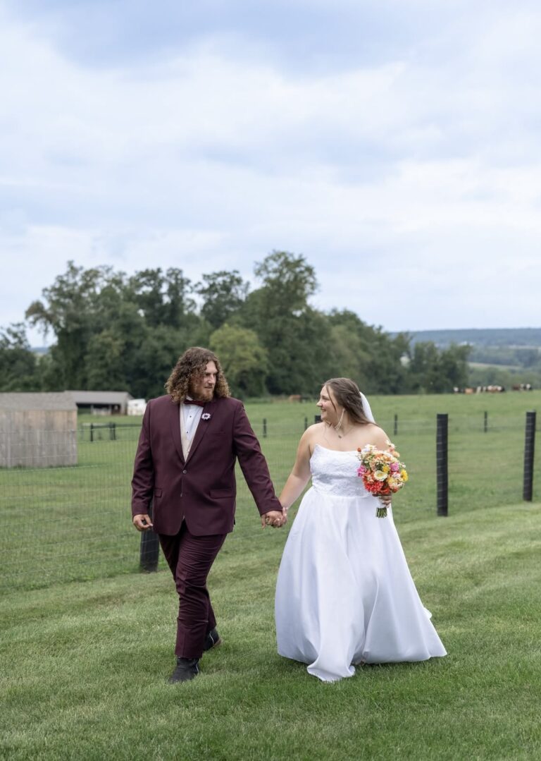 Bride in a white wedding gown and groom in a burgundy suit walk hand in hand across a grassy field with a fence in the background.