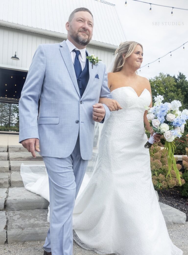 Bride in a white strapless wedding gown holding a blue-and-white bouquet walks down steps with a man in a light blue suit outdoors.
