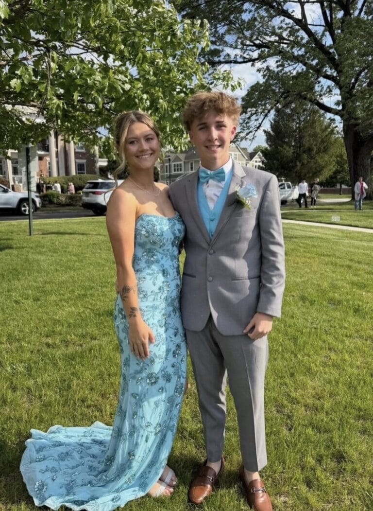 Young couple posing on a grassy lawn; woman in a teal strapless gown with lace, man in a gray suit with teal vest and bow tie.
