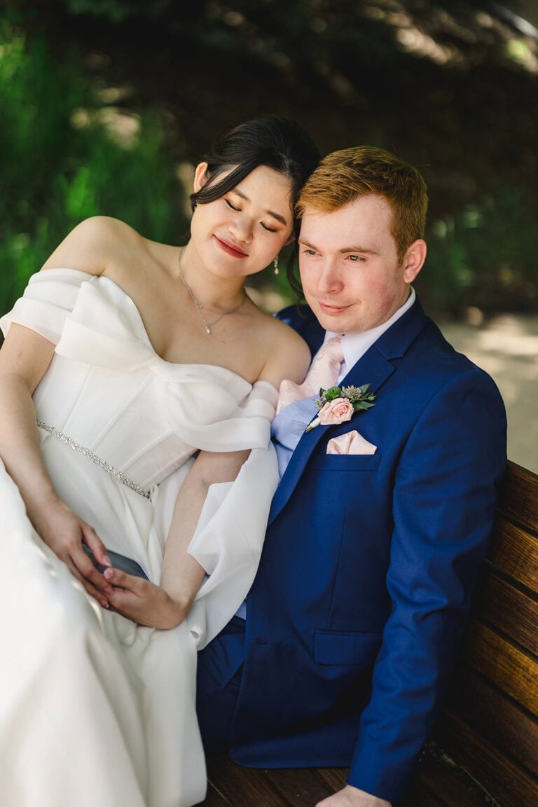 Jennifer Andrew Wedding Day | National Tuxedo Rentals Bride in an off-shoulder white wedding dress rests on a wooden bench beside her groom in a blue suit, both smiling softly with eyes closed.