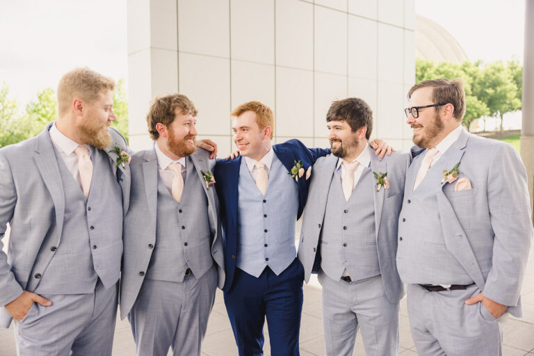 Five groomsmen in light gray suits with peach ties, arms around each other, smiling outdoors by a modern building. A man in a navy suit stands center-left as part of the group.