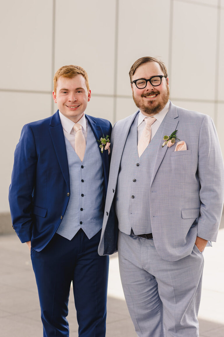 Two men in suits posing together; navy suit and light gray suit with pink ties and boutonnieres.