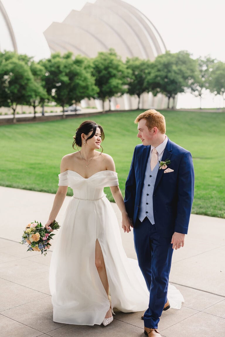 Jennifer Andrew Wedding Day | National Tuxedo Rentals Bride in a white off-shoulder gown and a blue-suited groom hold hands while walking outdoors with a bouquet of flowers.