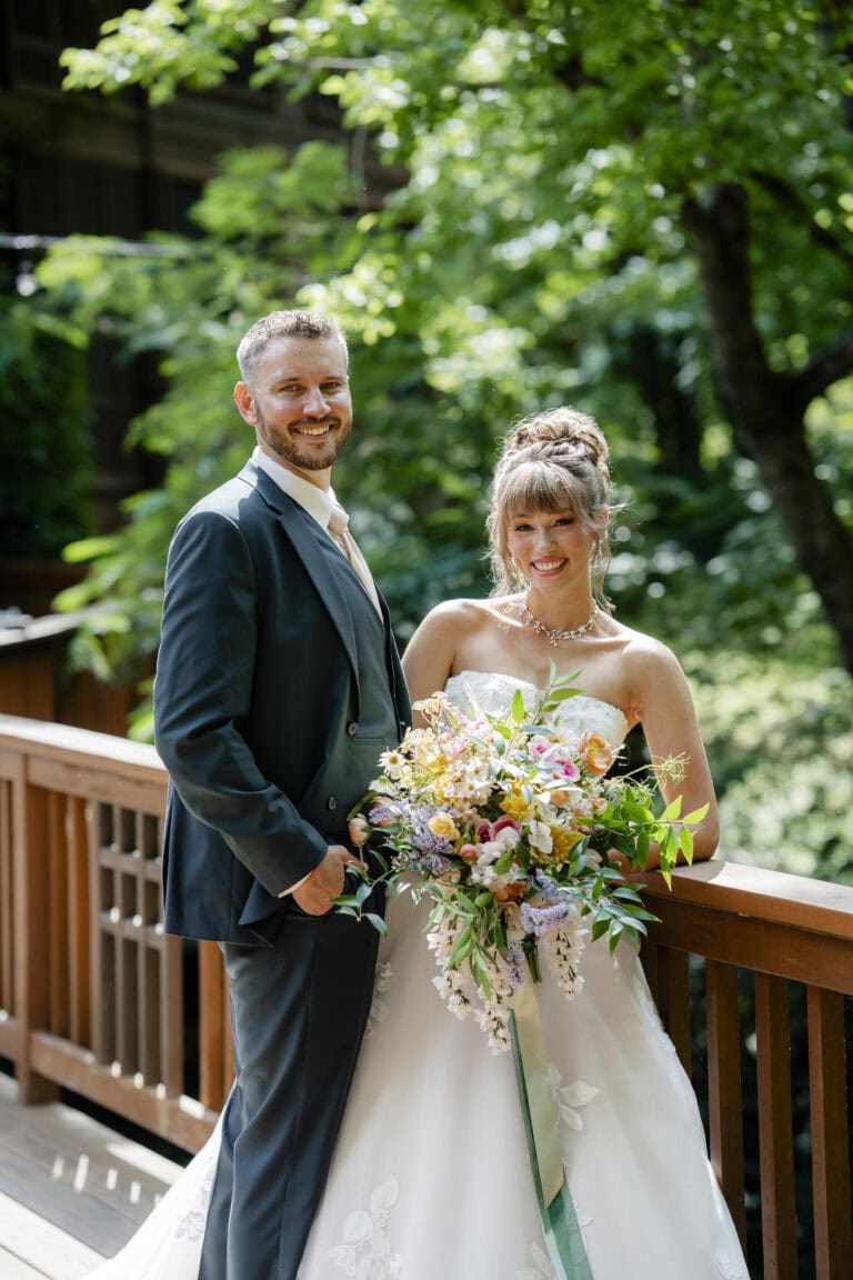MMWED-0376 | National Tuxedo Rentals Bride and groom pose together on a wooden railing outdoors, the bride holding a colorful bouquet and wearing a white wedding dress while the groom wears a navy suit and tie, smiling at the camera.