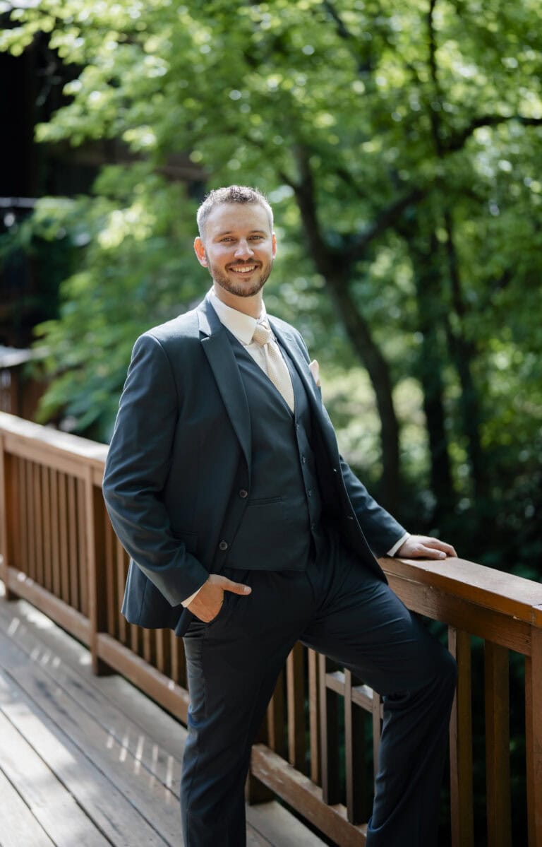 MMWED-0422 1 | National Tuxedo Rentals Smiling man in a navy suit leaning on a wooden railing outdoors among green trees.