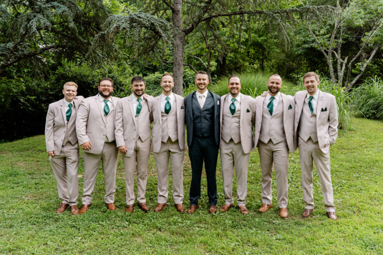 Group of seven men in beige suits with green ties posing outdoors in a garden setting.