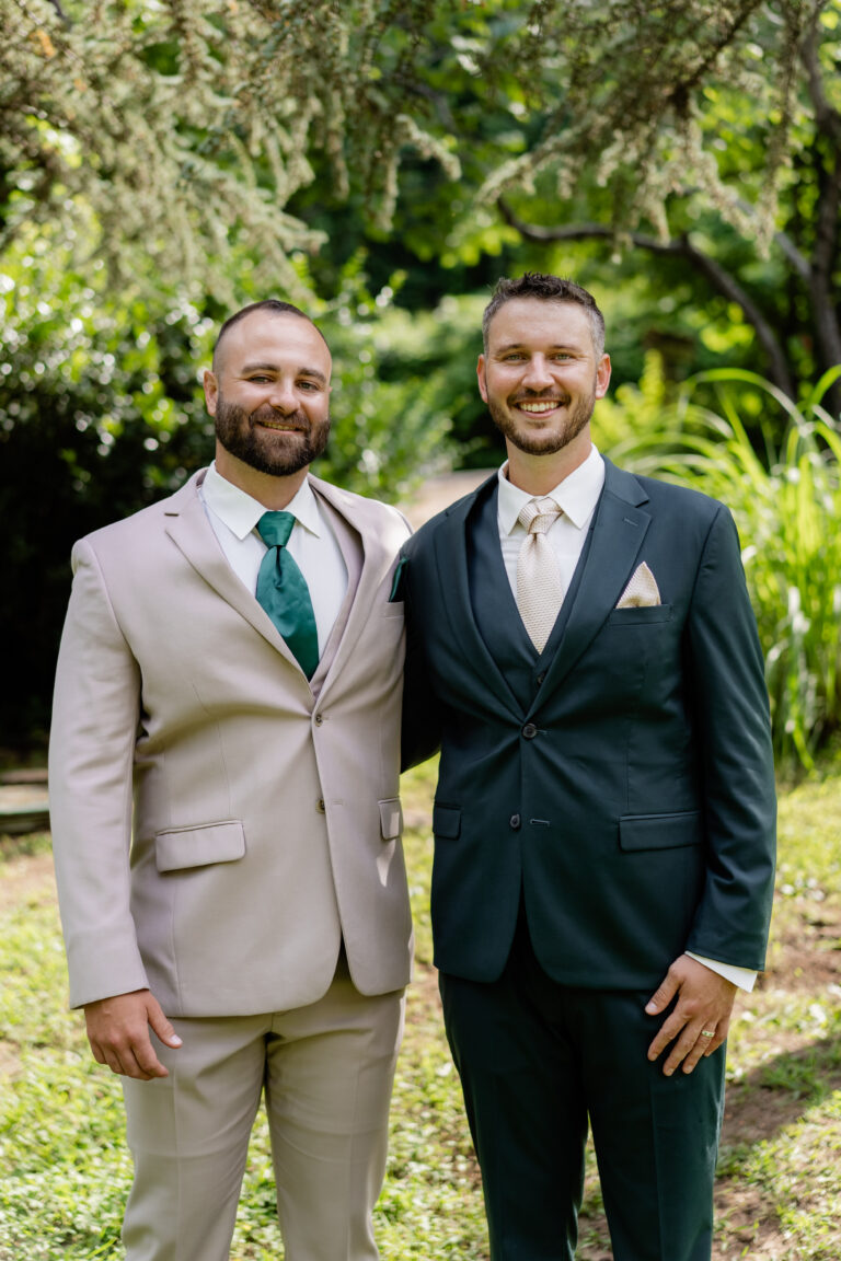 Two men in suits standing outdoors in a garden, smiling at the camera.