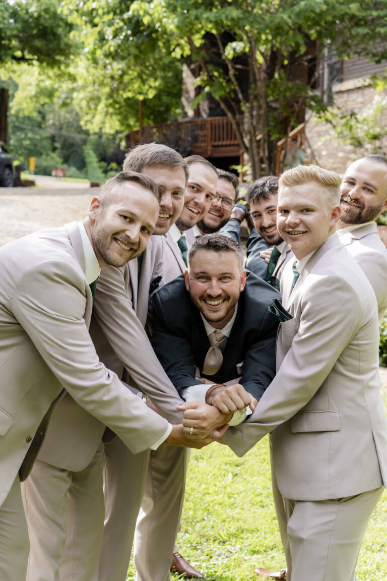 Groomsmen in beige suits join hands in a circle, smiling at the camera outdoors on a sunny lawn.