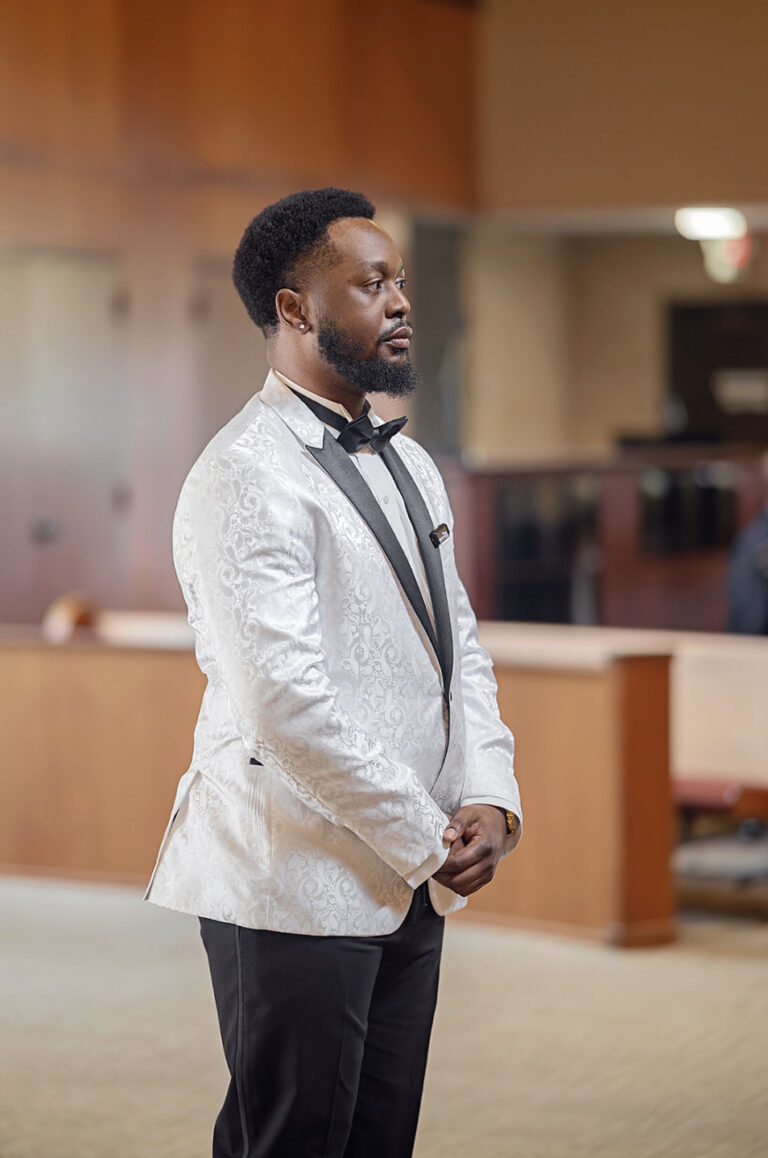 Formal-dressed man in a white brocade jacket and black bow tie stands in a lobby, hands clasped in front.