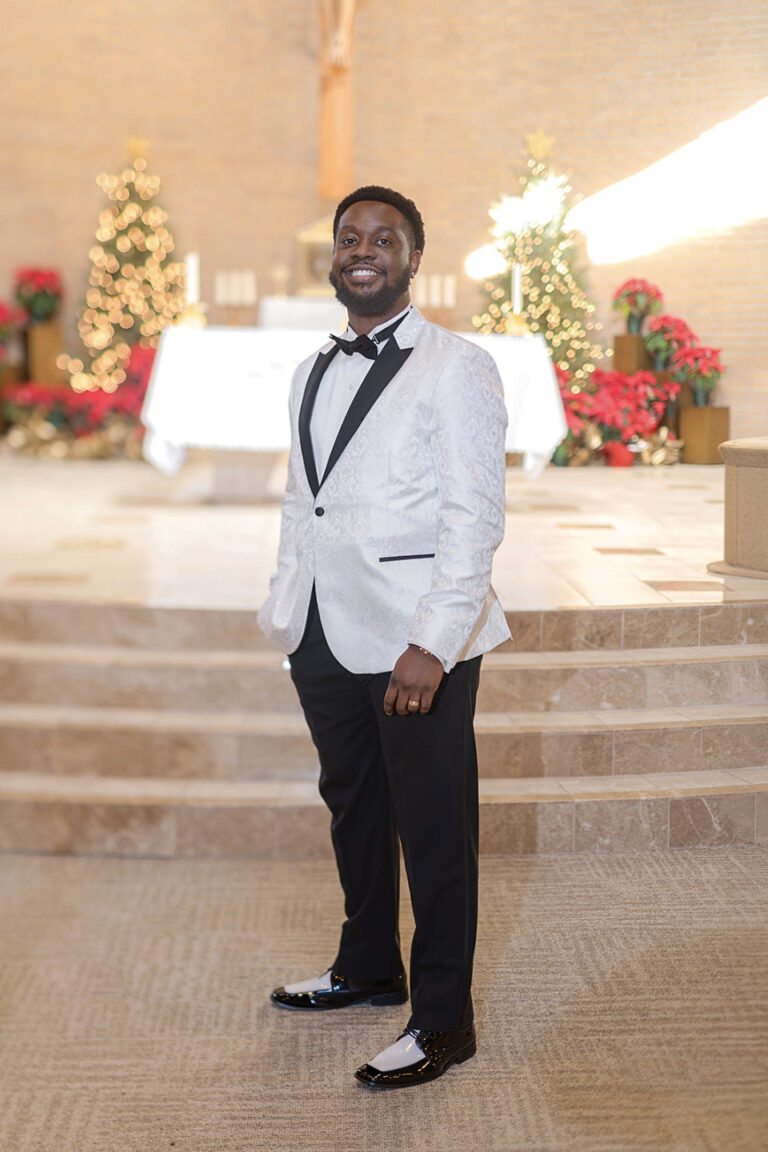 Man in a white patterned tuxedo jacket and black bow tie posing in a church hall with Christmas trees and poinsettias in the background.