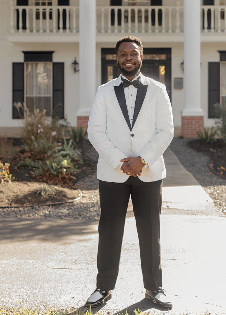 Man in a white tuxedo with black lapels and bow tie standing outdoors in front of a house with columns, smiling confidently.