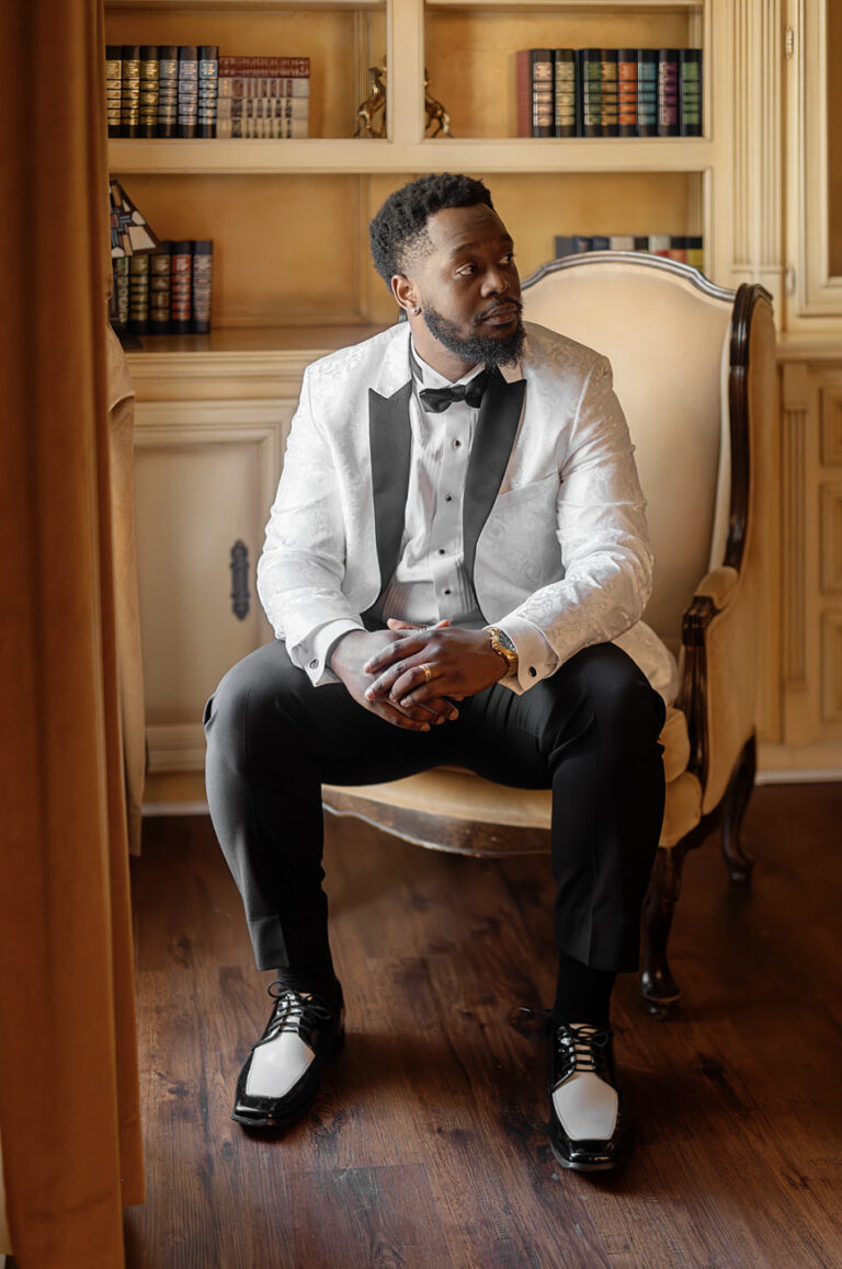 Man in a white tuxedo jacket and black bow tie sits in an elegant library, hands clasped, looking to the side.