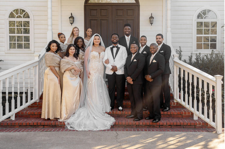Wedding party posing on brick steps in front of a white house; bride and groom center with bridesmaids in gold gowns and groomsmen in black suits nearby.