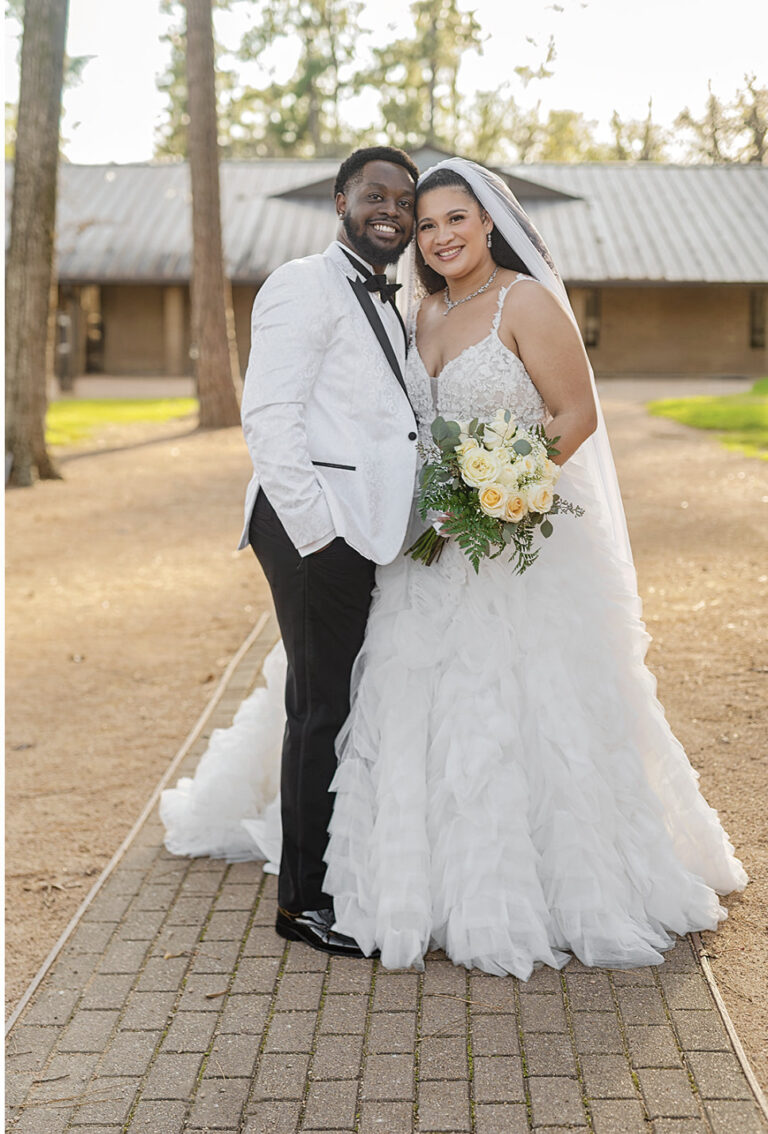 Bride and groom pose outdoors in wedding attire, the bride holding a bouquet of white roses.