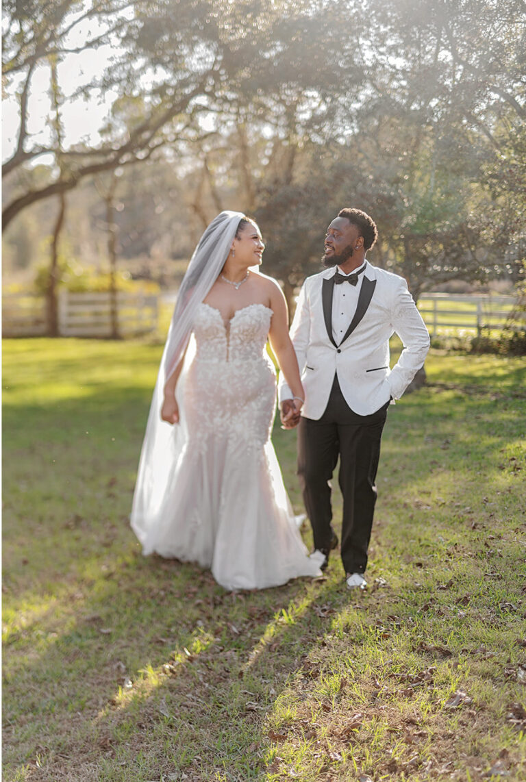 Bride and groom walk hand in hand outdoors, smiling, as sunlight filters through trees in a grassy field during a wedding.