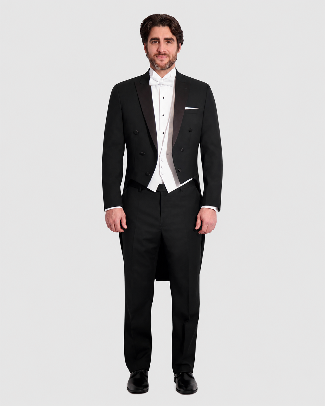 Man in a black tuxedo with a white waistcoat and bow tie, tails visible, standing against a light background.