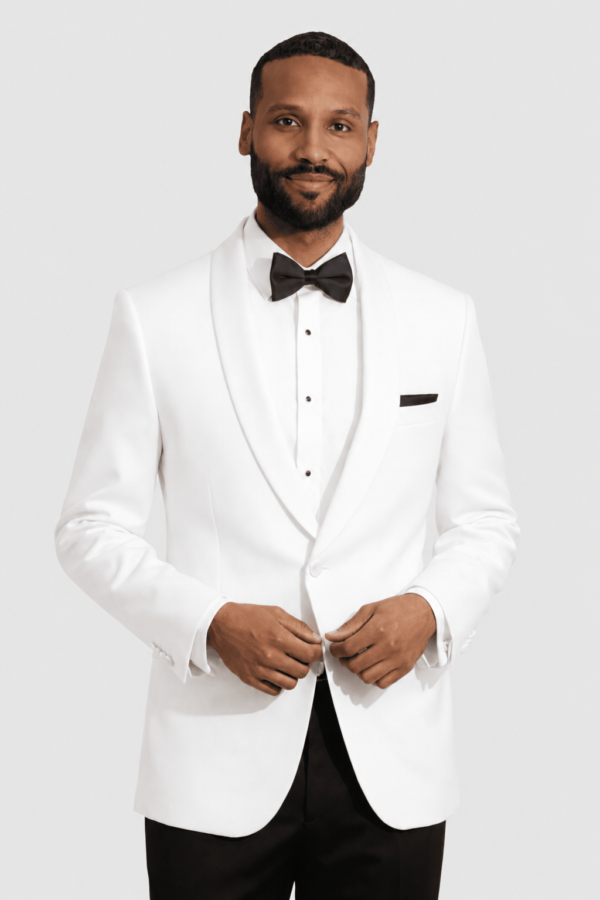 Man in a white tuxedo with a black bow tie, posing with hands clasped in front against a light background, smiling subtly.