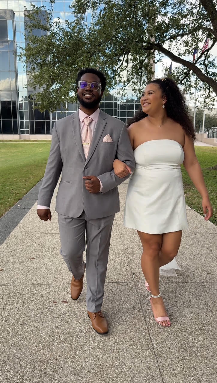 Couple walking arm-in-arm on a sunny sidewalk outside a glass-front building, dressed in formal attire and smiling at each other.