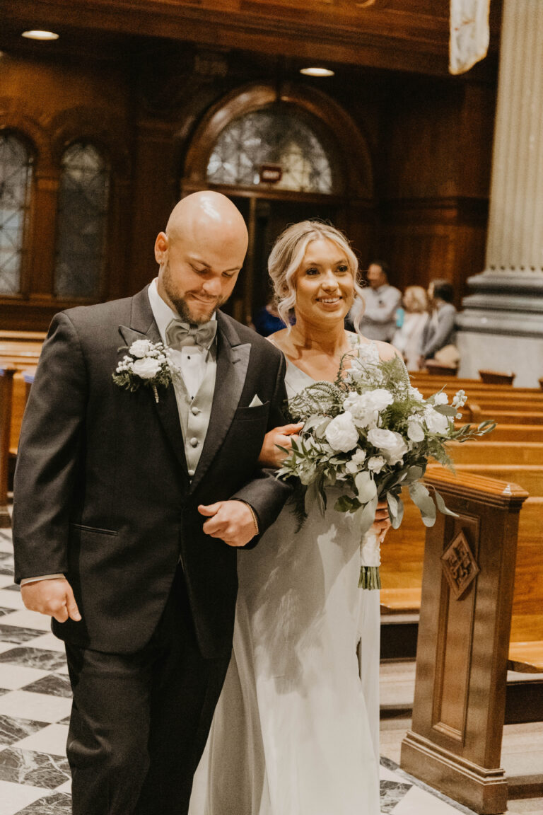 Bride and groom in formal attire walking arm in arm inside a church, the bride holding a white bouquet.